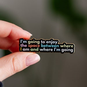 May include: Black enamel pin with gold trim featuring the text "I'm going to enjoy the space between where I am and where I'm going". The text is in rainbow colours.