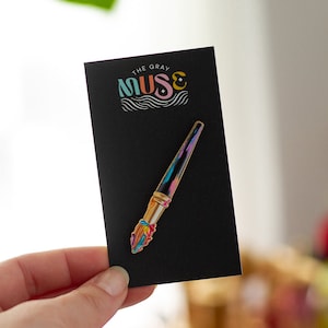 Puede incluir: Un pin de esmalte de pincel dorado con salpicaduras de pintura coloridas en las cerdas. El pin está en una tarjeta negra con el texto "THE GRAY MUSE" en letras de colores.