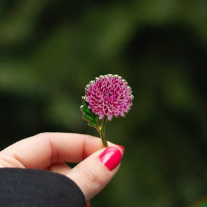 以下が含まれることがあります： 金色の仕上げが施されたピンクのエナメル製のフラワーピン。花は丸く、細かい花びらが特徴です。ピンはピンクのネイルを塗った手で持たれています。