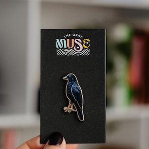 Puede incluir: Un pin de esmalte negro con un pájaro azul oscuro con detalles dorados, montado en una tarjeta negra. La tarjeta tiene el texto "THE GRAY MUSE" en letras coloridas encima de la ilustración del pájaro. El pin es sostenido por una persona.