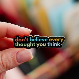 May include: A rectangular enamel pin with the text "don't believe every thought you think" in colorful lettering. The pin has a black border and is held by a person's hand.