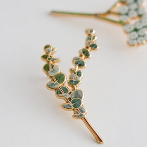May include: A gold-coloured metal eucalyptus leaf pin with green and grey enamel detailing. The pin features a detailed design of eucalyptus leaves and stems. Other floral pins are visible in the background.