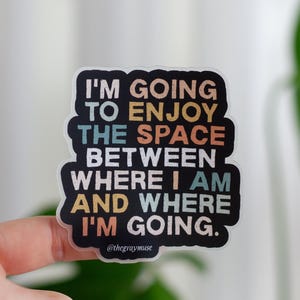 May include: A sticker with a black background and colorful text that reads "I'm going to enjoy the space between where I am and where I'm going." The text is in a rainbow color scheme and the sticker has a white border.
