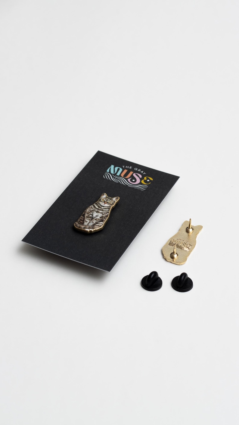 May include: A gold-colored enamel pin featuring a tabby cat sitting with a black background. The pin is packaged on a black card with the text "The Gray Muse" in colorful lettering.