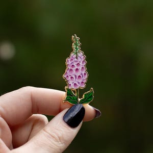 May include: A gold-tone enamel pin featuring a pink and green foxglove flower.