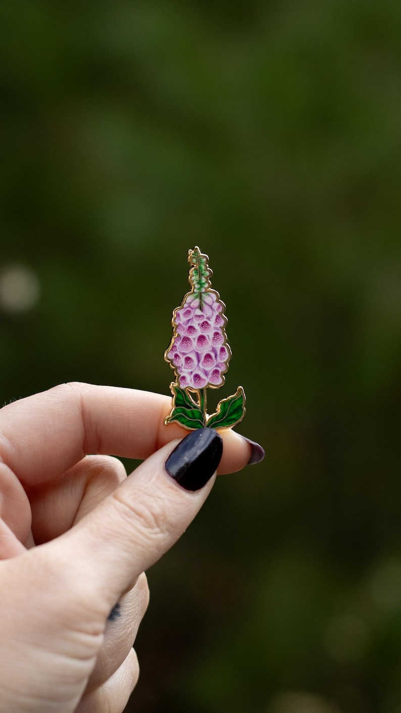 May include: A gold-tone enamel pin featuring a pink and green foxglove flower.