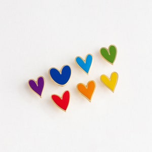 May include: Seven heart-shaped pins in a rainbow of colors. The pins are gold-toned with a red, orange, yellow, green, blue, indigo, and violet enamel fill.