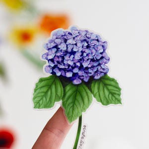 Puede incluir: Una pegatina de flor de hortensia morada con hojas verdes y tallo. La pegatina est&aacute; en un dedo y tiene el texto "@thegraymuse" en el tallo.