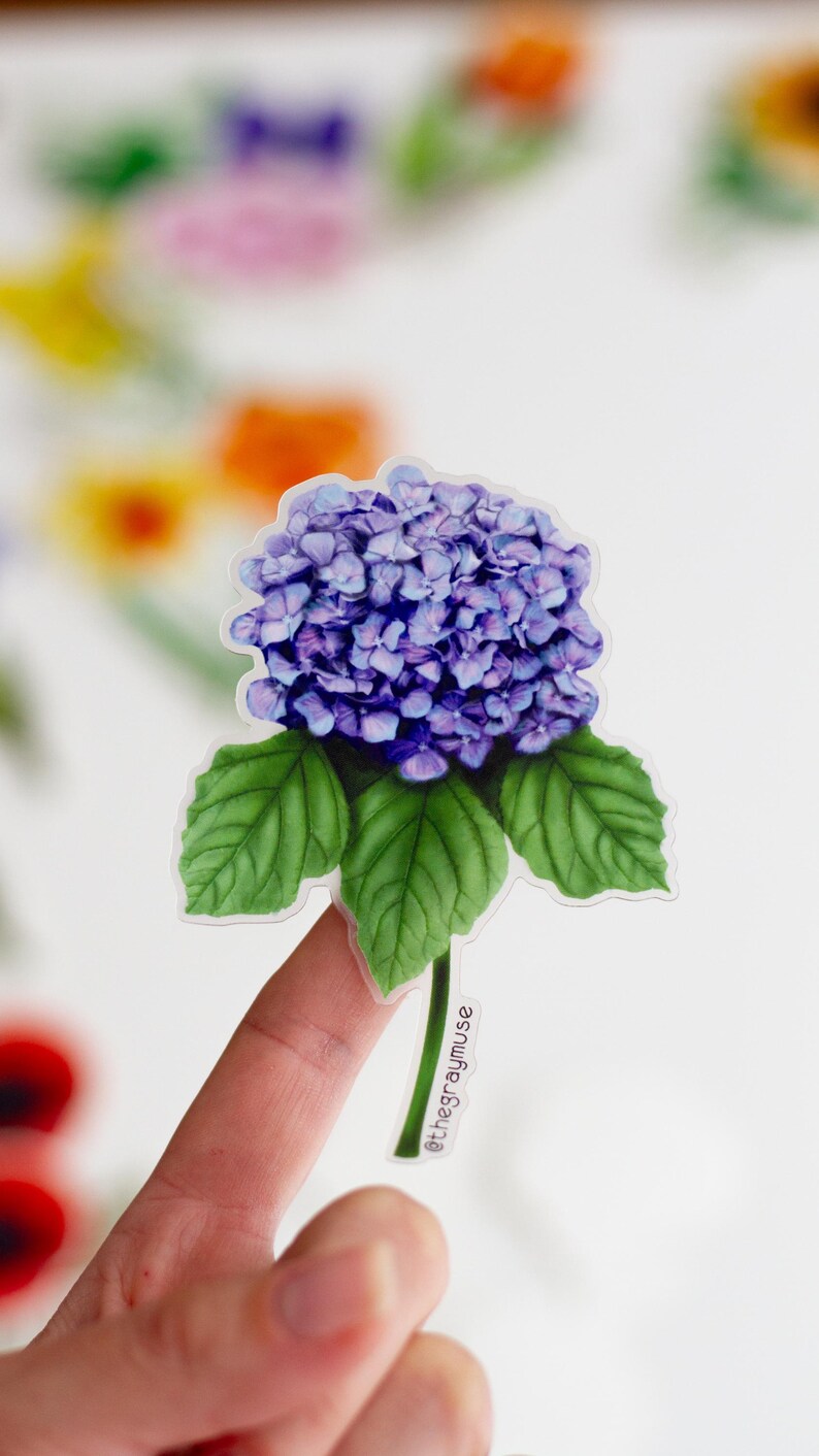 Puede incluir: Una pegatina de flor de hortensia morada con hojas verdes y tallo. La pegatina est&aacute; en un dedo y tiene el texto "@thegraymuse" en el tallo.