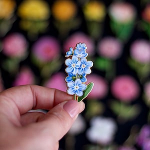 May include: A gold-toned enamel pin featuring a cluster of blue forget-me-not flowers with green leaves.