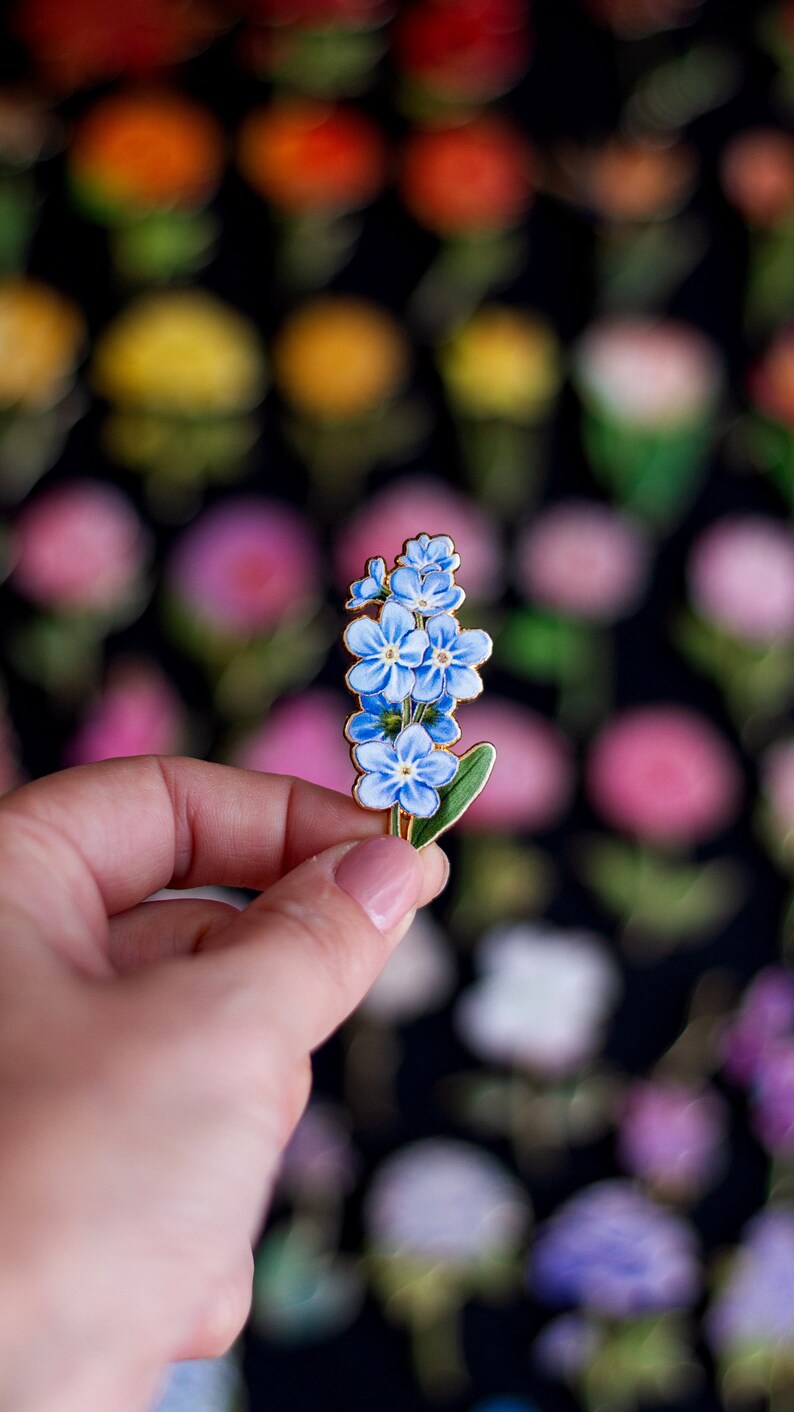 May include: A gold-toned enamel pin featuring a cluster of blue forget-me-not flowers with green leaves.