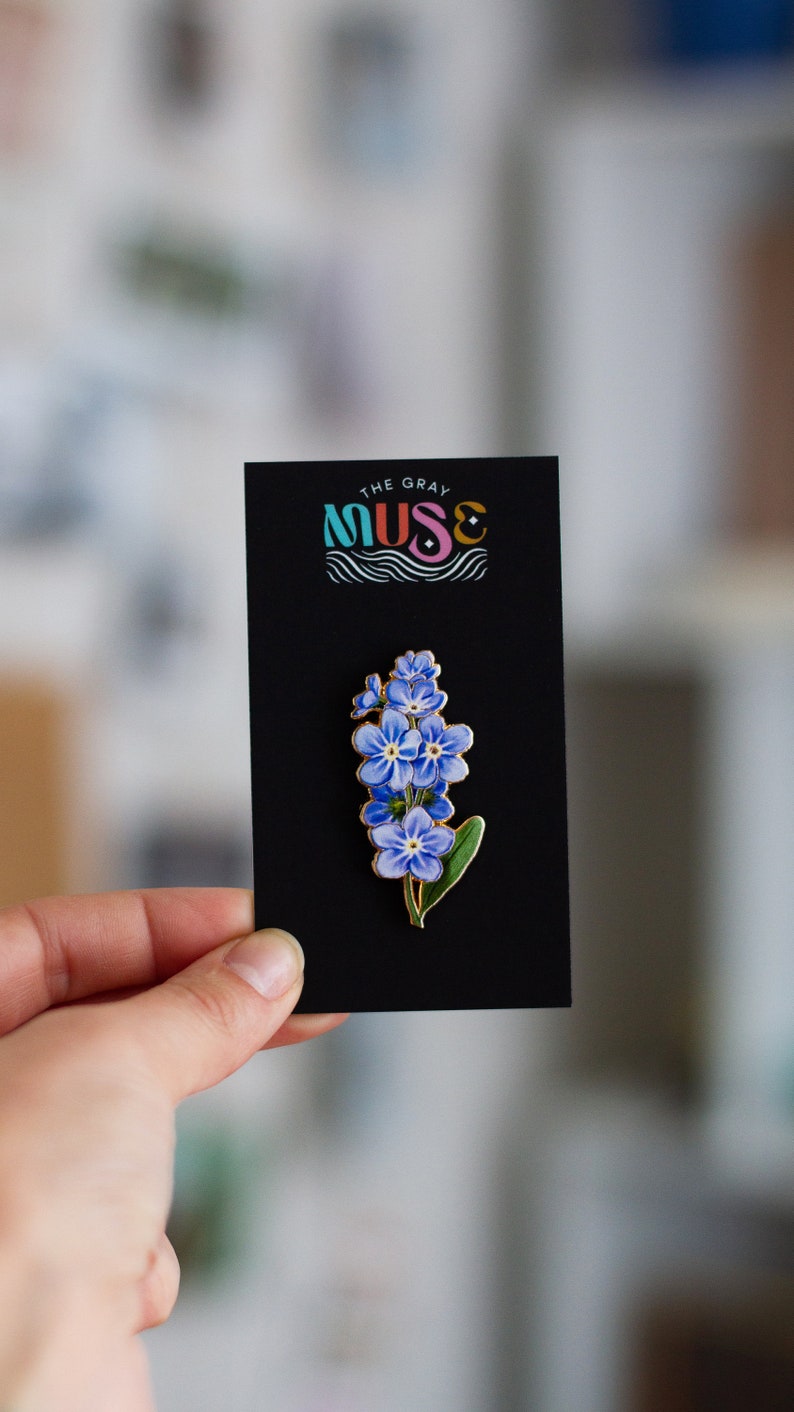 May include: A black card with the text "The Gray Muse" in colorful letters and a blue and green wave design. A gold enamel pin with a cluster of blue forget-me-not flowers is displayed on the card.