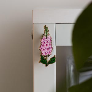 May include: A pink and green enamel flower pin with a gold finish. The flower is a stylized foxglove.