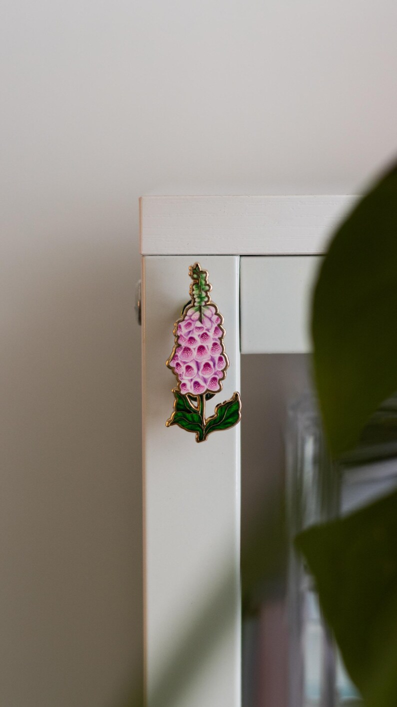 May include: A pink and green enamel flower pin with a gold finish. The flower is a stylized foxglove.