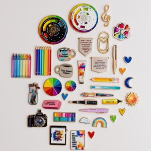 May include: A collection of art-related pins and accessories, such as paint palettes, pencils, and a camera, arranged on a white surface. The image includes pins with text like "Not Paint Water" and "My brain has 100 tabs open."