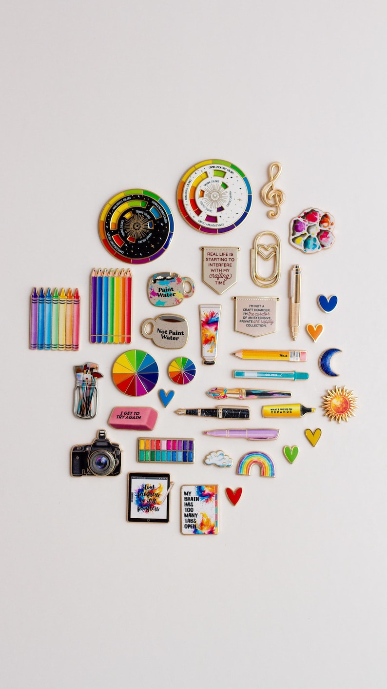 May include: A collection of art-related pins and accessories, such as paint palettes, pencils, and a camera, arranged on a white surface. The image includes pins with text like "Not Paint Water" and "My brain has 100 tabs open."