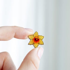 May include: A yellow daffodil flower pin with a red and orange center. The pin is held between two fingers against a white background. The flower has a gold-colored outline.