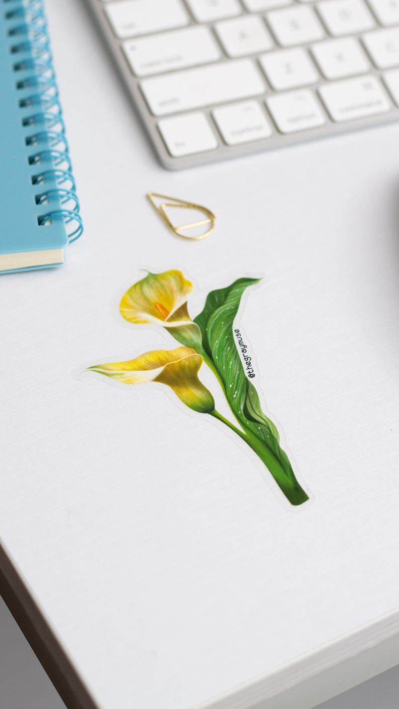 May include: A sticker featuring a detailed illustration of two yellow calla lilies with green leaves. The sticker has a white border and the text "@thegreymuse" is visible on the green leaf. The sticker is on a white surface.