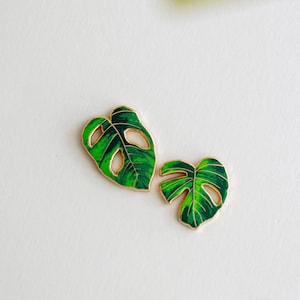 May include: A pair of gold-rimmed enamel earrings shaped like monstera leaves. The leaves feature vibrant green gradients with dark green veins and distinct cut-out holes, capturing the natural appearance of the plant foliage in a small accessory.