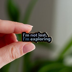 May include: A black enamel pin with gold trim featuring the text "I'm not lost, I'm exploring".