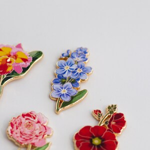 May include: A set of five enamel flower pins. The pins feature a variety of flowers, including a red poppy, a pink peony, a blue forget-me-not, and a yellow orchid.