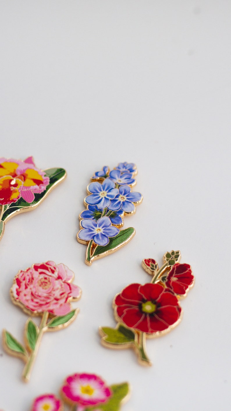 May include: A set of five enamel flower pins. The pins feature a variety of flowers, including a red poppy, a pink peony, a blue forget-me-not, and a yellow orchid.