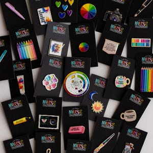 May include: A selection of enamel pins displayed on black cards. The pins showcase diverse designs, such as a rainbow, a camera, a pencil, a paintbrush, and a colour wheel. The pins are vibrant and detailed, with a range of shapes and sizes.