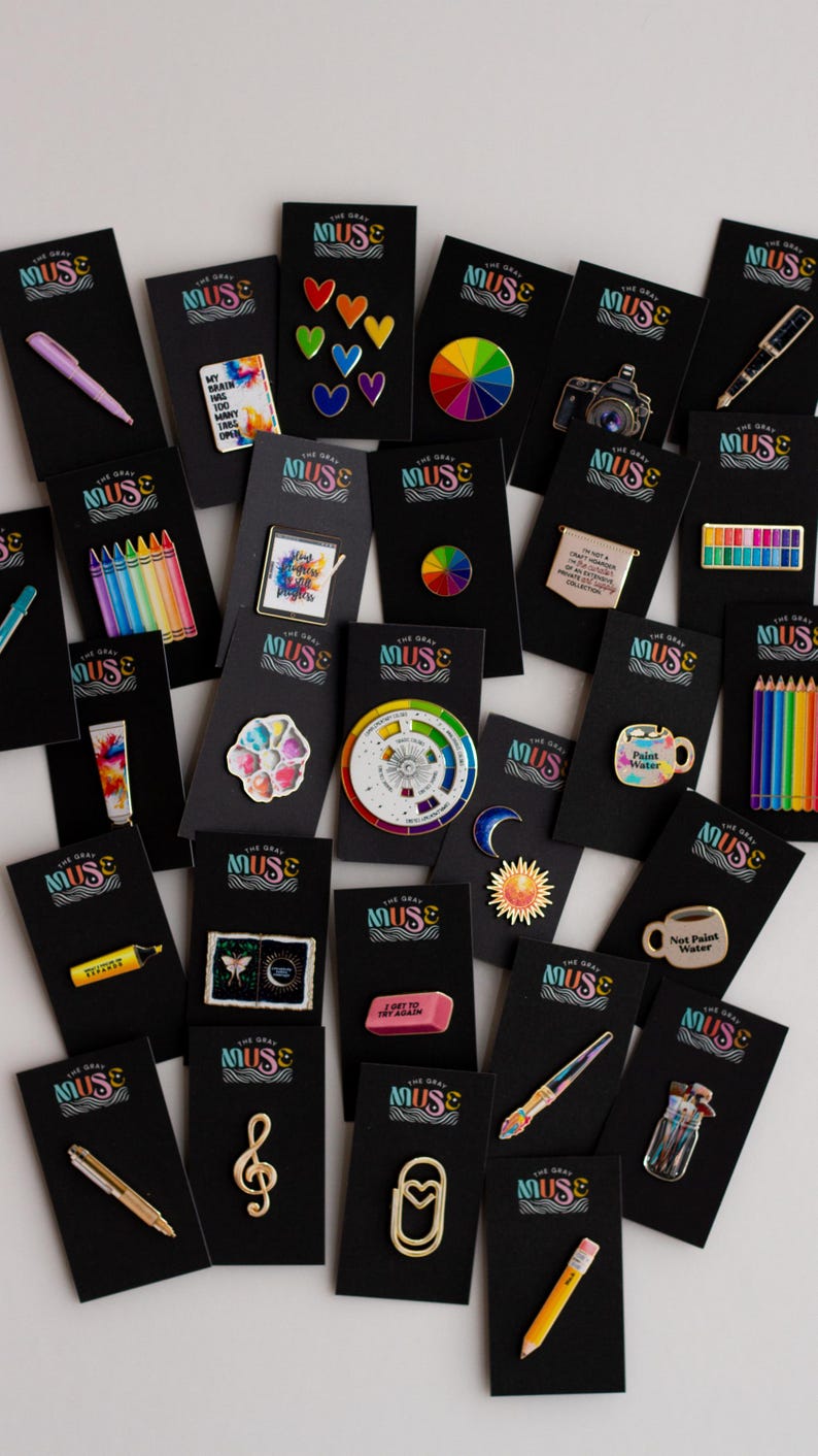 May include: A selection of enamel pins displayed on black cards. The pins showcase diverse designs, such as a rainbow, a camera, a pencil, a paintbrush, and a colour wheel. The pins are vibrant and detailed, with a range of shapes and sizes.