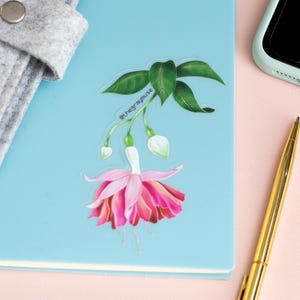 May include: A clear sticker with a pink fuchsia flower with green leaves and the text "@thegraymuse" on a blue notebook.