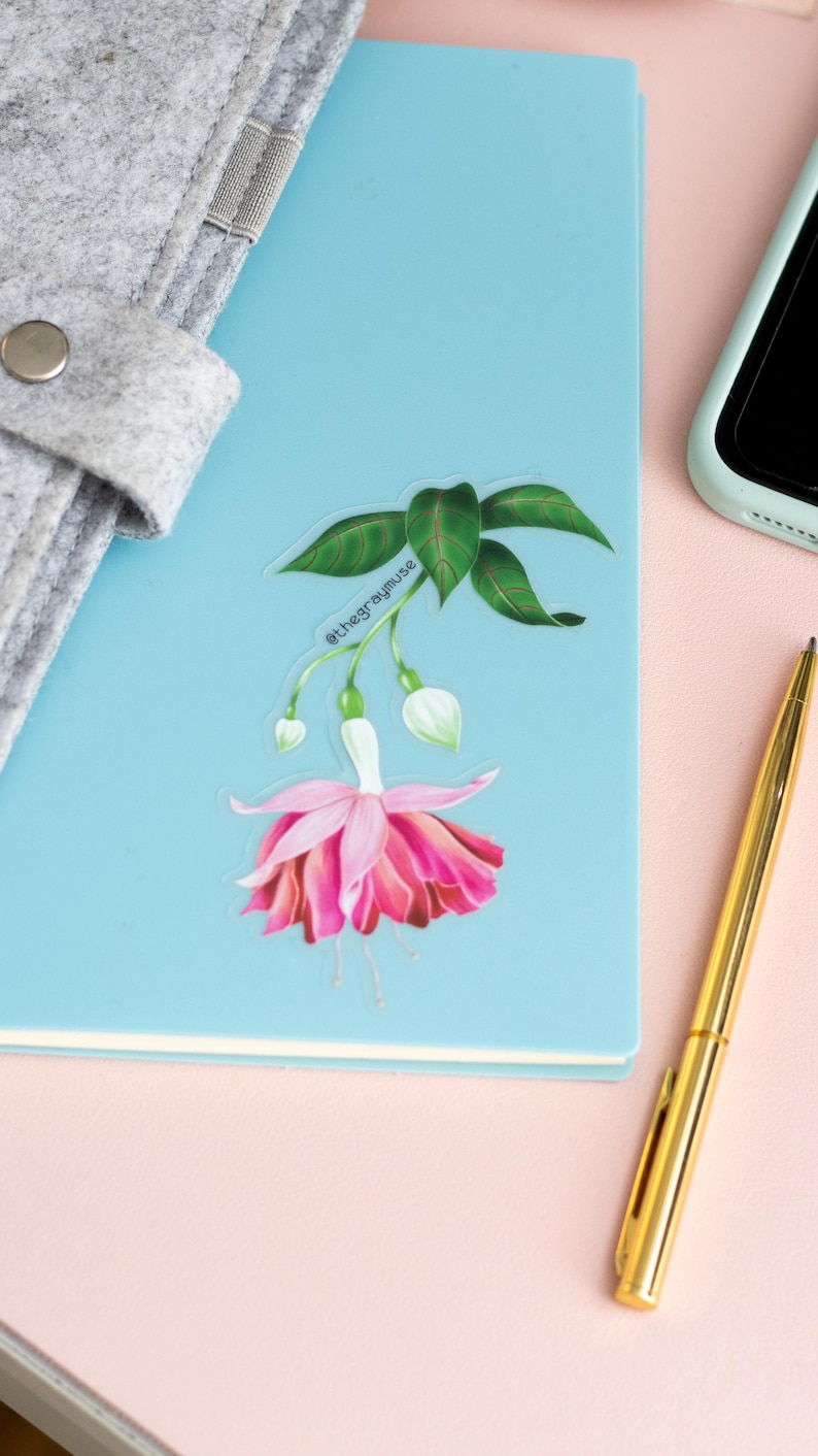 May include: A clear sticker with a pink fuchsia flower with green leaves and the text "@thegraymuse" on a blue notebook.