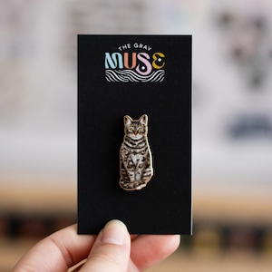May include: A gold-colored enamel pin featuring a tabby cat sitting with a black background. The pin has the text "The Gray Muse" in colorful letters above the cat.