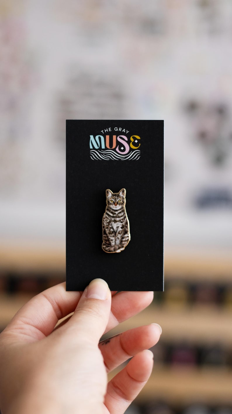 May include: A gold-colored enamel pin featuring a tabby cat sitting with a black background. The pin has the text "The Gray Muse" in colorful letters above the cat.