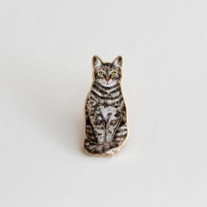 May include: A gold-toned pin featuring a detailed illustration of a tabby cat sitting with its paws tucked in. The cat has brown and white stripes and yellow eyes.