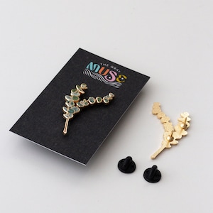 May include: A gold-coloured metal pin in the shape of a sprig of eucalyptus leaves with blue-green accents. The pin is on a black card with the text "THE GRAY MUSE" in a colourful design. Two black pin backs are also visible.