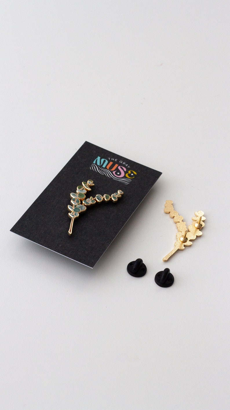 May include: A gold-coloured metal pin in the shape of a sprig of eucalyptus leaves with blue-green accents. The pin is on a black card with the text "THE GRAY MUSE" in a colourful design. Two black pin backs are also visible.