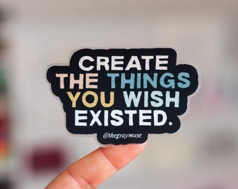 May include: A clear sticker with black text that reads "CREATE THE THINGS YOU WISH EXISTED."  The text is in a rainbow gradient of colors. The sticker is held by a hand with pink nail polish. The text at the bottom of the sticker reads "@thegraymuse".