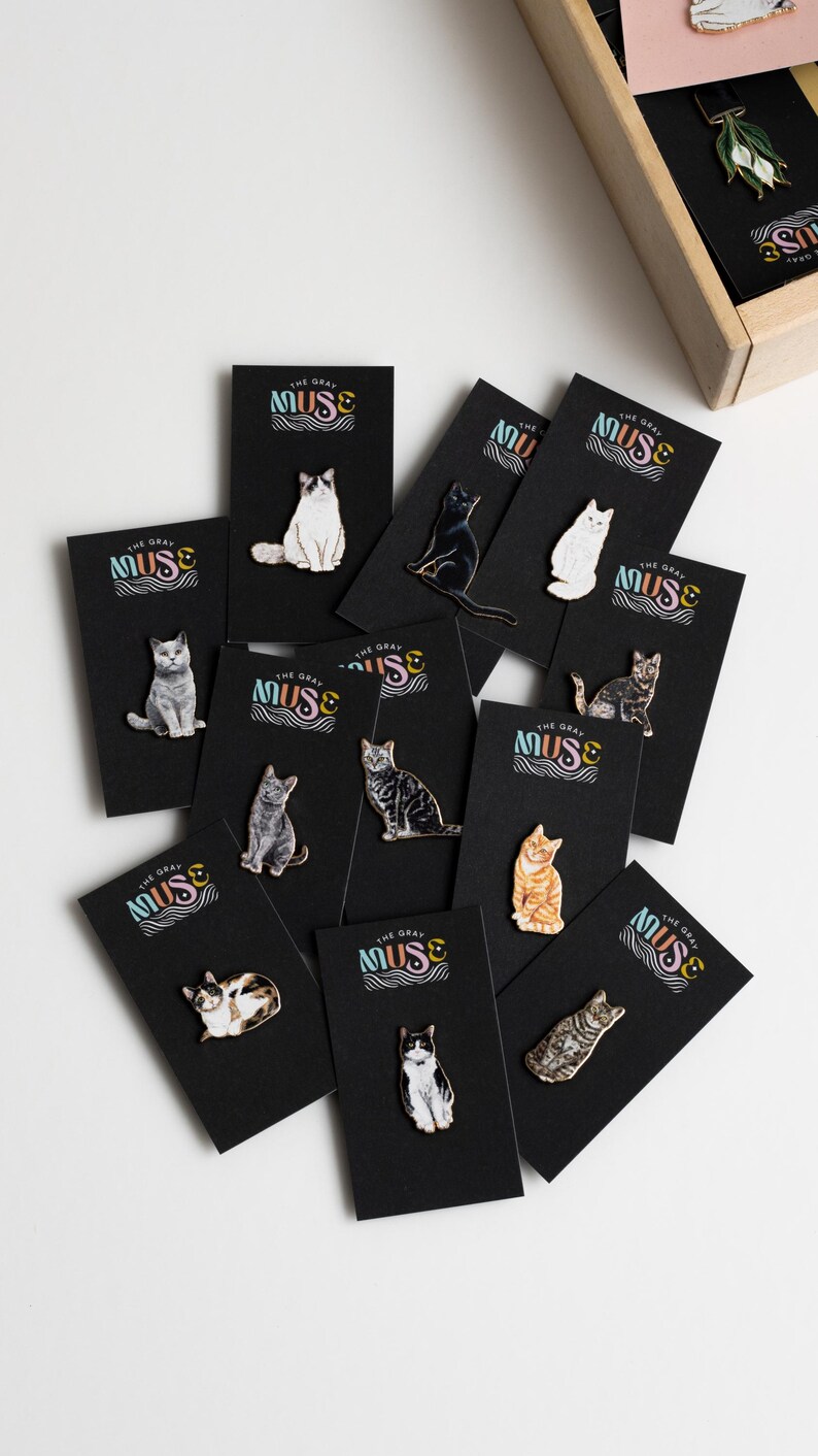 May include: A collection of 10 enamel pins featuring different cat breeds. Each pin is on a black card with the text "The Gray Muse" in colorful letters. The pins are all different colors and have a glossy finish.