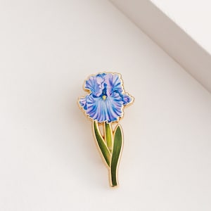 May include: A gold-toned enamel pin featuring a purple iris flower with green leaves.