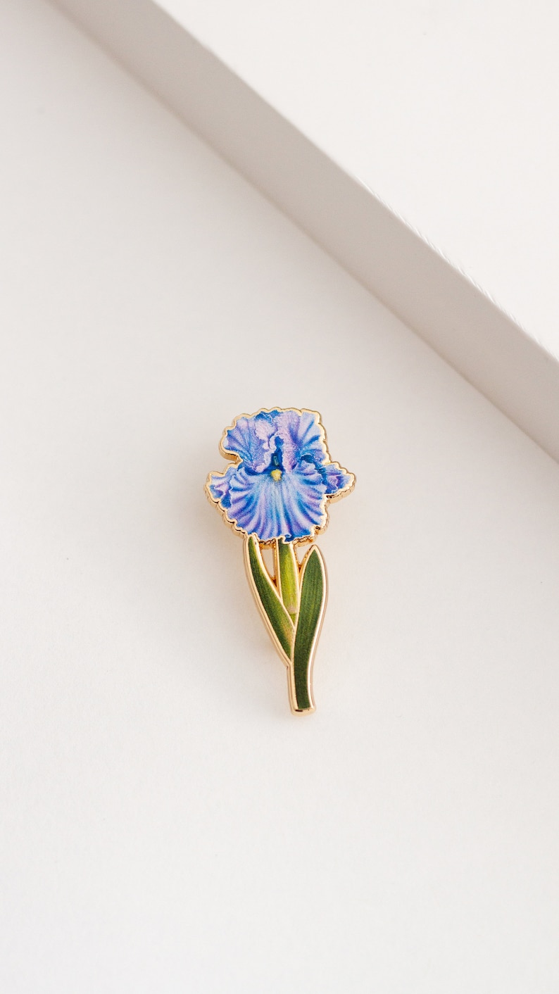 May include: A gold-toned enamel pin featuring a purple iris flower with green leaves.