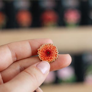 May include: A small, round pin featuring a detailed orange and red flower design with a gold border and a green center. The pin is held between two fingers, showcasing its intricate floral pattern and vibrant colors.