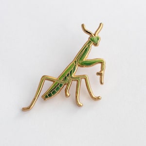 May include: A gold-toned metal pin in the shape of a praying mantis. The insect has a green body with black detailing and gold-colored legs and antennae. The pin is a decorative accessory.