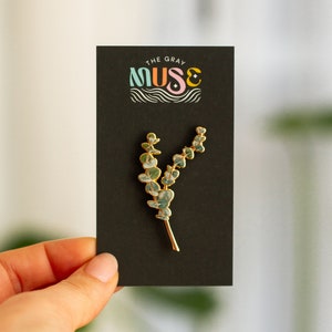 May include: A gold-coloured metal pin shaped like a eucalyptus branch, featuring green and blue enamel leaves. The pin is presented on a black card with the text "THE GRAY MUSE" in colourful lettering.