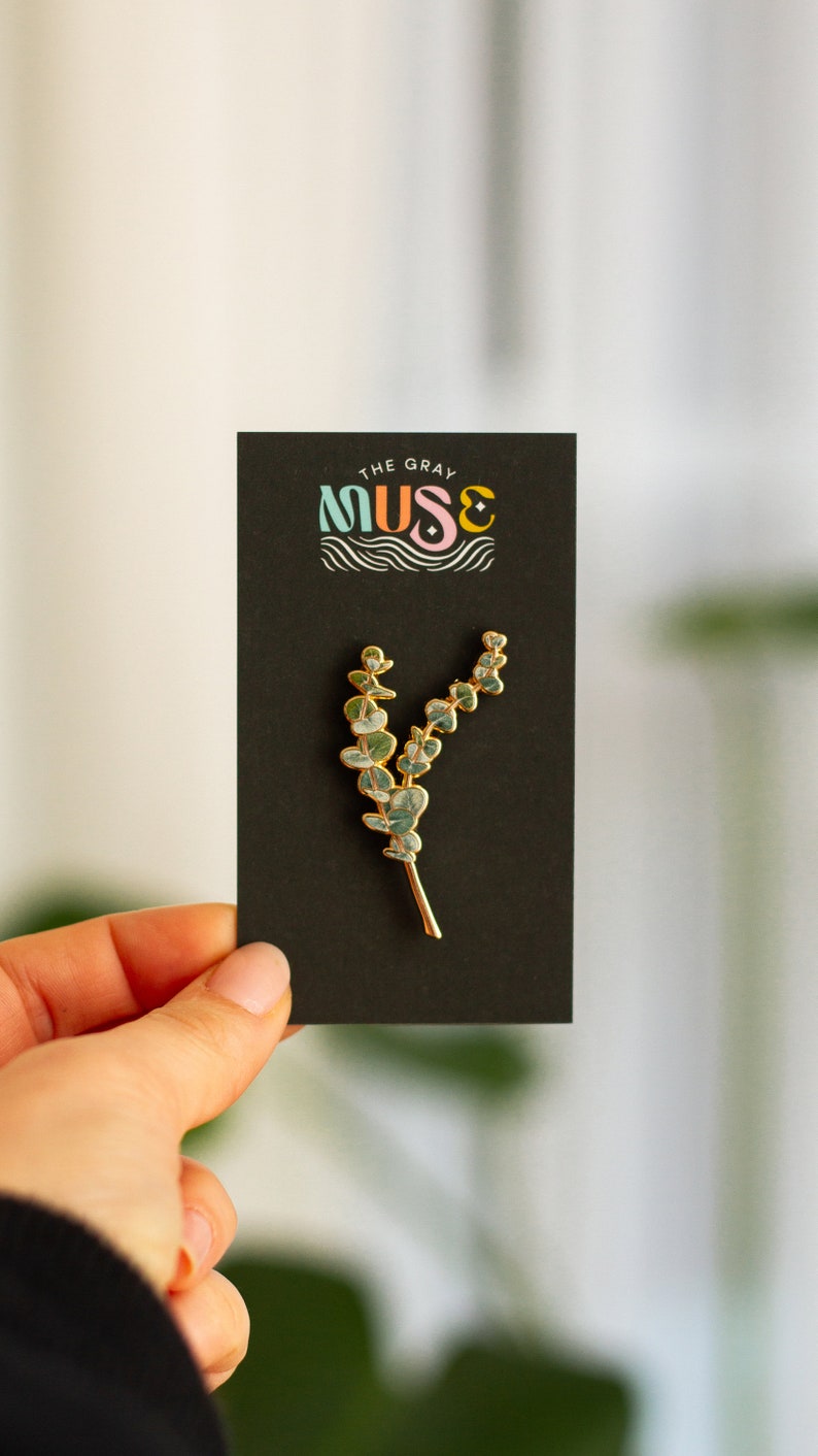 May include: A gold-coloured metal pin shaped like a eucalyptus branch, featuring green and blue enamel leaves. The pin is presented on a black card with the text "THE GRAY MUSE" in colourful lettering.
