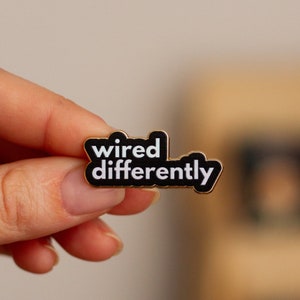 以下が含まれることがあります： 黒のエナメルピンで、白い文字で「wired differently」と書かれています。