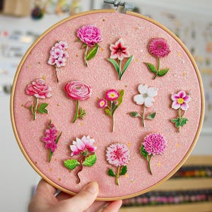 K&ouml;nnte beinhalten: Ein rosa glitzernder Stickrahmen mit verschiedenen floralen Designs. Die Blumen sind in Rosa- und Wei&szlig;t&ouml;nen gehalten, mit gr&uuml;nen Stielen und Bl&auml;ttern. Der Rahmen wird von einer Hand gehalten, die das Handwerk und Design zeigt.