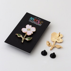 K&ouml;nnte beinhalten: Ein bl&uuml;tenf&ouml;rmiger Emaille-Anstecker mit rosa Bl&uuml;tenbl&auml;ttern, gr&uuml;nen Bl&auml;ttern und goldfarbenen Akzenten. Der Anstecker wird auf einer schwarzen Karte mit dem Text "MUSE THE GRAY" pr&auml;sentiert. Zwei schwarze Pin-R&uuml;ckseiten sind ebenfalls sichtbar.