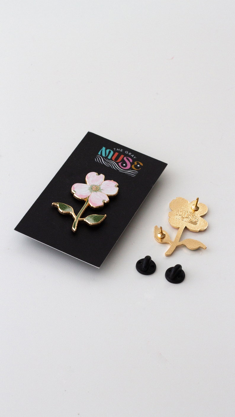 K&ouml;nnte beinhalten: Ein bl&uuml;tenf&ouml;rmiger Emaille-Anstecker mit rosa Bl&uuml;tenbl&auml;ttern, gr&uuml;nen Bl&auml;ttern und goldfarbenen Akzenten. Der Anstecker wird auf einer schwarzen Karte mit dem Text "MUSE THE GRAY" pr&auml;sentiert. Zwei schwarze Pin-R&uuml;ckseiten sind ebenfalls sichtbar.