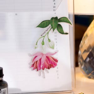May include: A pink fuchsia flower sticker with green leaves and the text "@thegraymuse" on a white lined paper background.