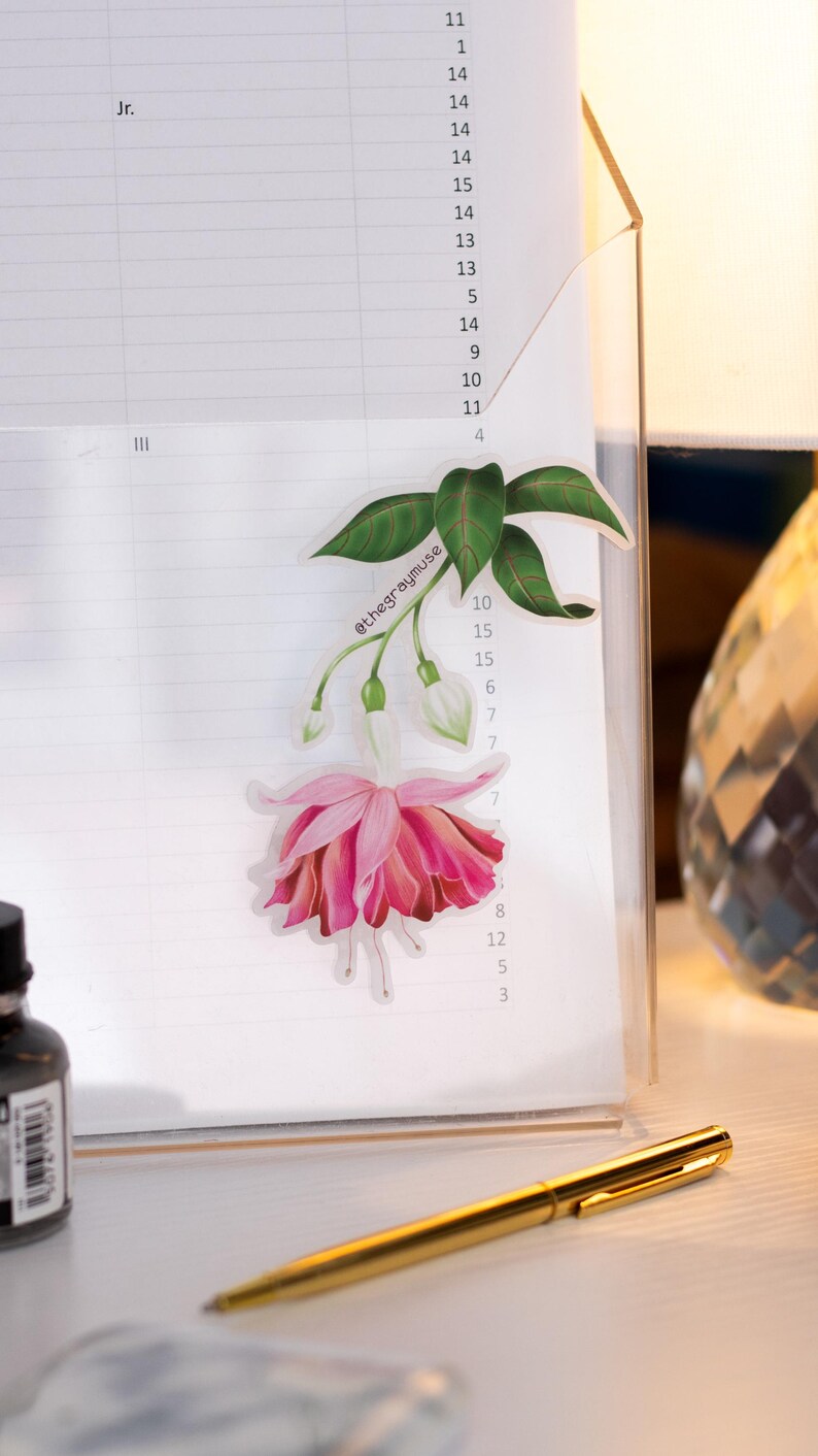 May include: A pink fuchsia flower sticker with green leaves and the text "@thegraymuse" on a white lined paper background.