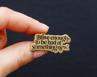 May include: Gold-colored enamel pin with the text "Brave enough to be bad at something new."